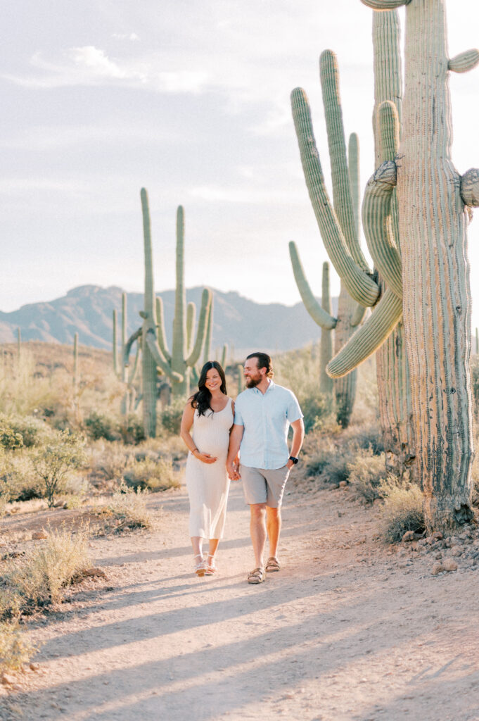 Tucson Maternity Outdoor Session of a  couple walks hand in hand on a desert path surrounded by tall cacti. The woman is pregnant and wearing a white dress, while the man wears a light blue shirt and shorts. Mountains are visible in the background under a clear sky. 
