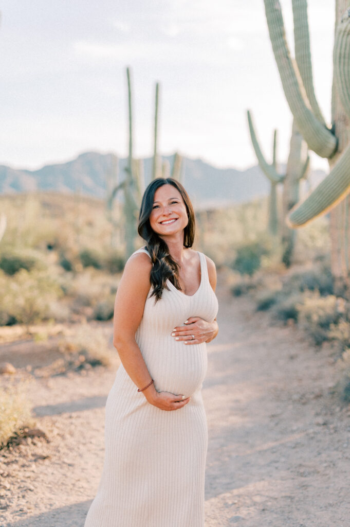 Tucson Maternity Outdoor Session of a smiling pregnant woman in a light, sleeveless dress stands on a desert trail surrounded by cacti, with mountains in the background under a clear sky. 