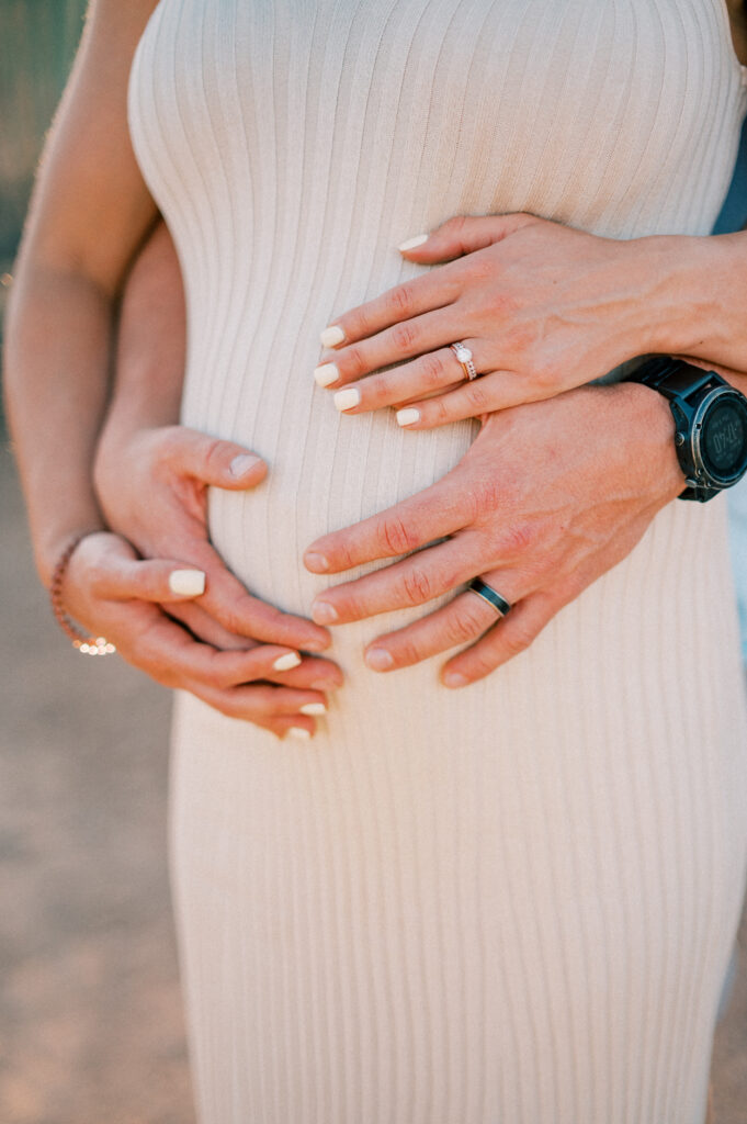  Close-up of a pregnant woman in a ribbed beige dress, with her hands and a mans hands gently resting on her baby bump, both wearing rings and having manicured nails. 
