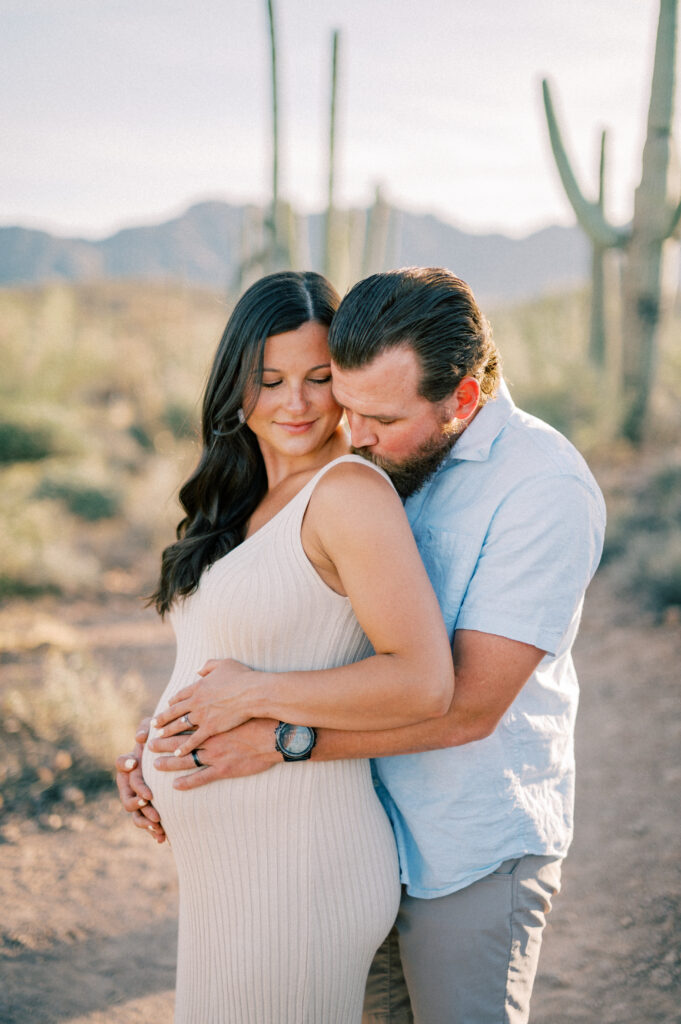  A man hugs a pregnant woman from behind on a desert trail with cacti and mountains in the background. Both appear happy and serene, with the mans hands gently resting on the womans baby bump. 