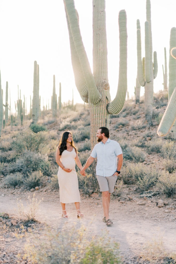Tucson Maternity Outdoor Session of a couple holds hands while walking on a desert path surrounded by tall cacti. The woman, who appears pregnant, wears a light dress, and the man wears a light shirt and shorts. The sun is bright in the background. 