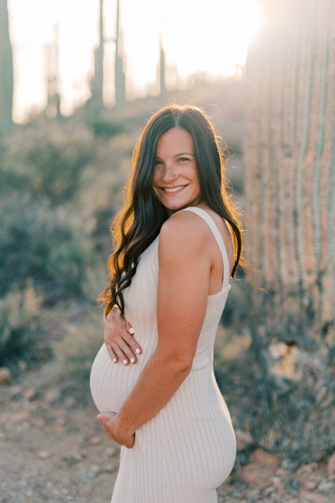  A woman in a sleeveless beige dress stands outdoors in sunlight, gently holding her pregnant belly and smiling at the camera, with desert plants and cacti in the background. 