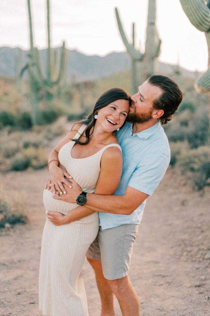  A smiling couple stands on a desert path with cacti in the background. The woman, visibly pregnant in a beige dress, is embraced from behind by the man in a light blue shirt and shorts, who kisses her on the head. 