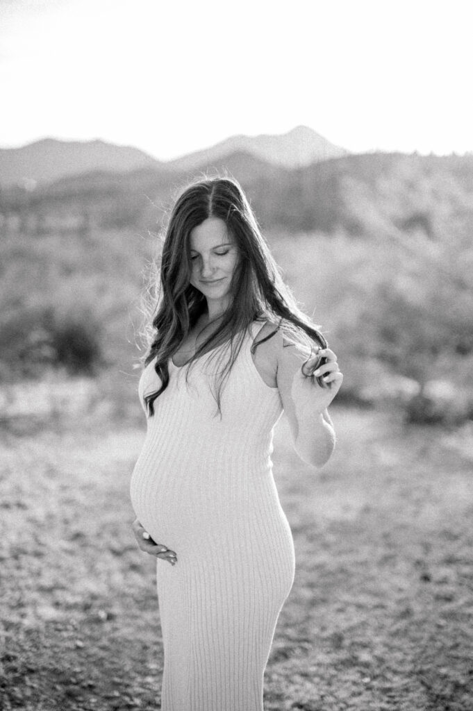 Tucson Maternity Outdoor Session of a pregnant woman in a long, sleeveless dress stands outdoors, gently holding her hair with one hand and resting the other on her belly. The background shows blurred mountains and natural scenery in soft light. 