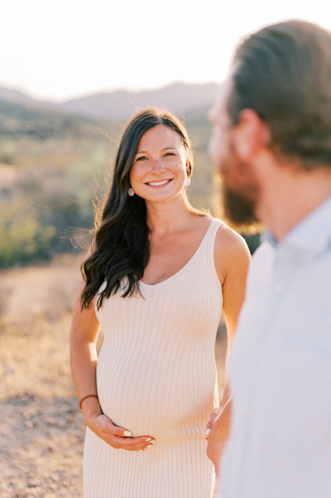  A pregnant woman in a light-colored dress smiles at the camera while standing outdoors, gently holding her belly. A bearded man in a light shirt stands beside her, slightly out of focus, with a natural landscape in the background. 