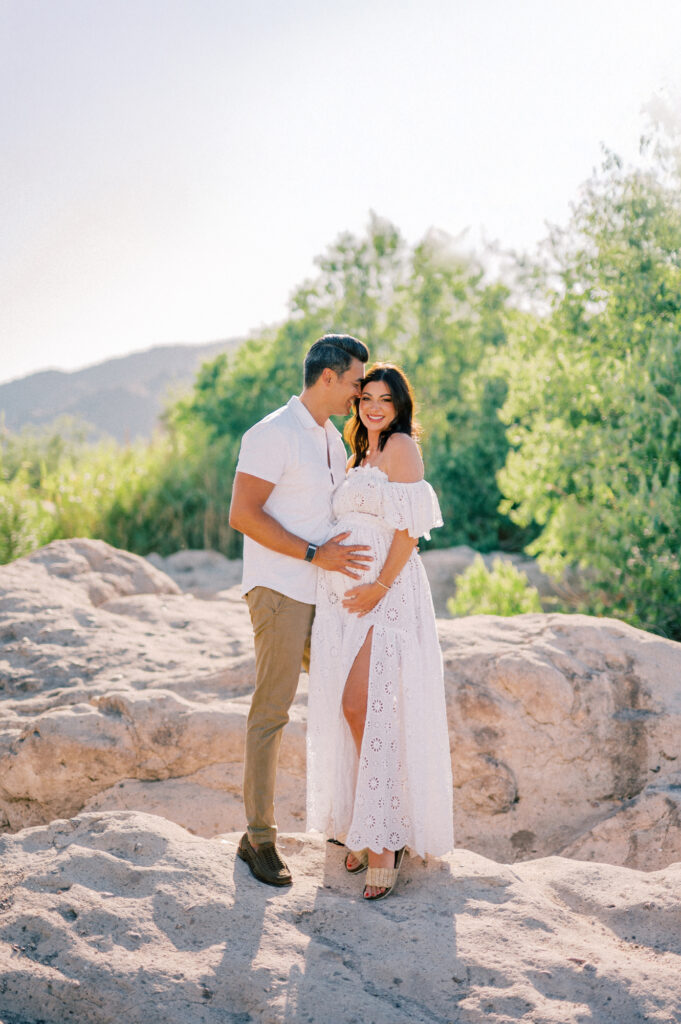 A couple stands outdoors on rocks for their Salt River Maternity shoot. The woman, in a white off-shoulder dress, holds her baby bump and smiles as her partner kisses her forehead. Lush green trees and hills complete the scenic backdrop.