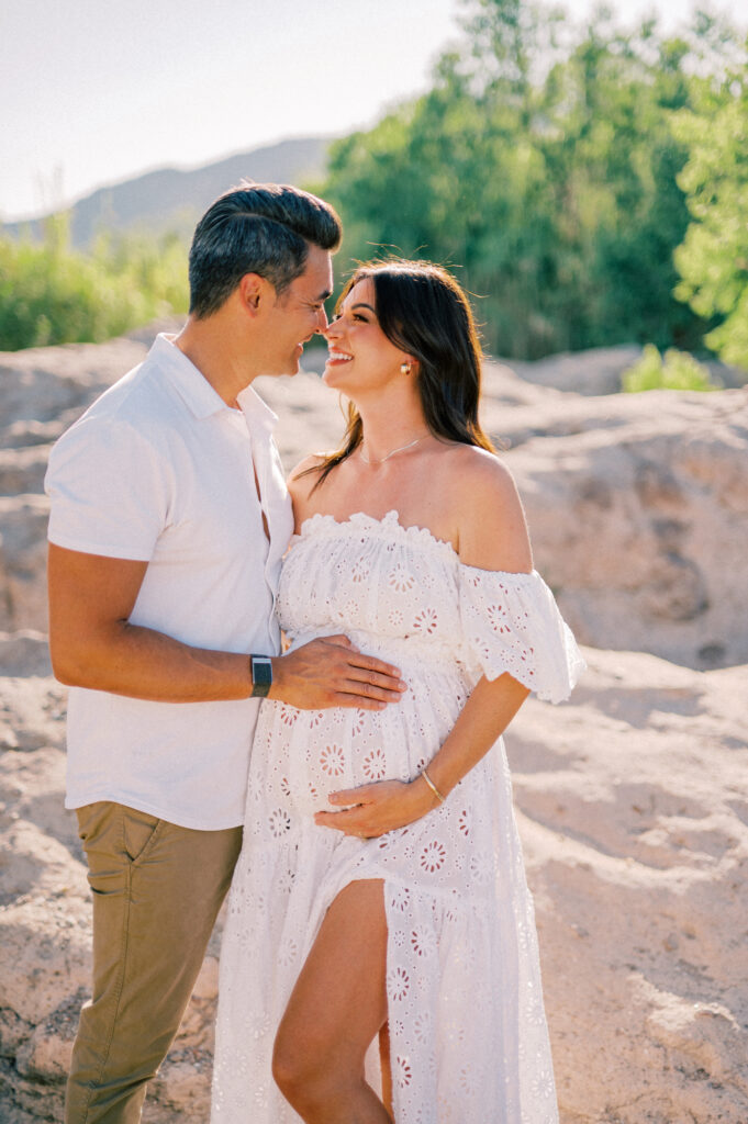 A couple stands outdoors on rocky terrain near the Salt River, smiling at each other. The woman, in a white off-the-shoulder dress, cradles her pregnant belly while he gently touches her bump. Lush greenery forms the perfect maternity backdrop.