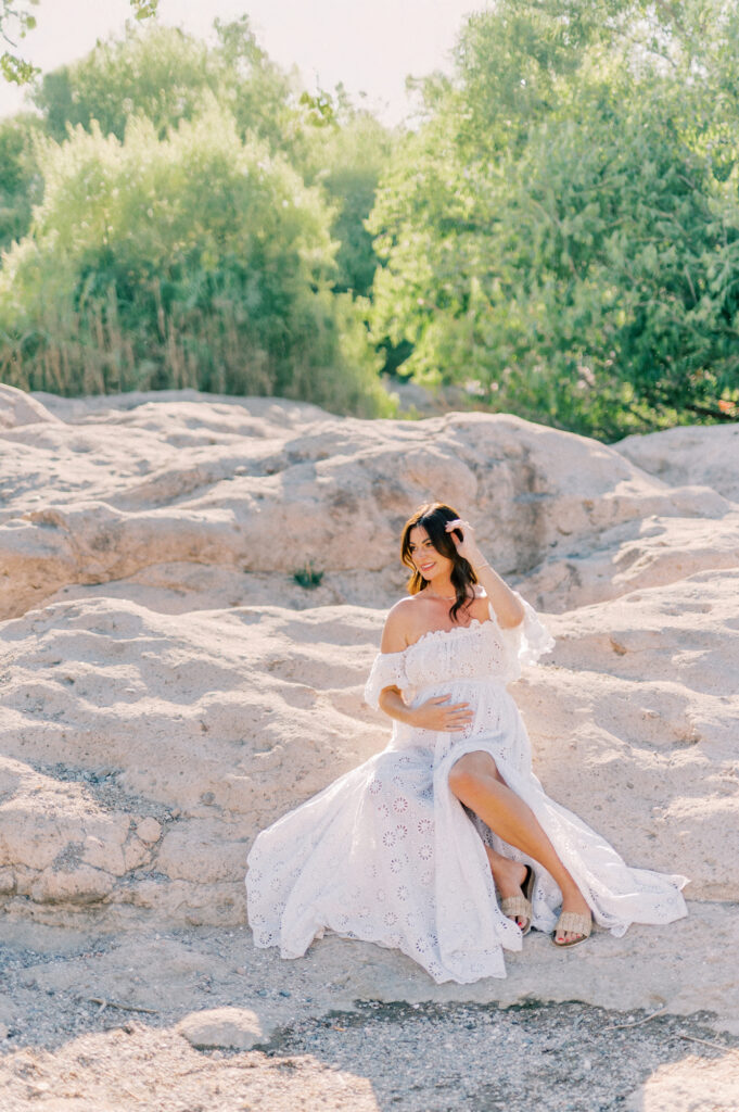 A woman in a flowing white dress sits on light-colored rocky terrain by the Salt River, surrounded by greenery and bathed in soft natural sunlight. She touches her hair and gazes to the side, creating a serene and elegant maternity scene.