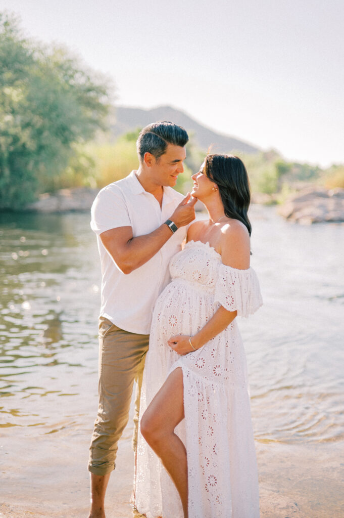 A couple stands by the Salt River, the man gently touching the woman's chin. The woman, visibly pregnant and wearing a white lace dress, cradles her belly. They look at each other lovingly, surrounded by nature and sunlight—a beautiful Salt River maternity moment.
