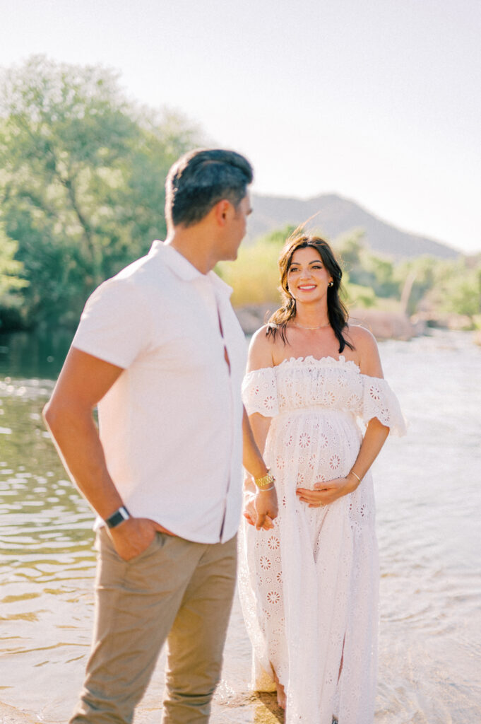 A pregnant woman in a white off-shoulder dress stands in shallow water, smiling and holding her belly. A man in a white shirt and khaki pants stands nearby, looking at her. Trees and hills are visible in the background.