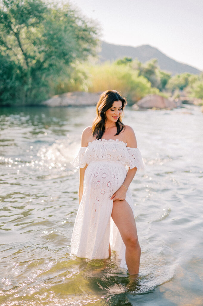 A pregnant woman in a white off-shoulder dress stands in shallow water, smiling and looking to the side. Green trees and distant hills are visible under a clear sky in the background.