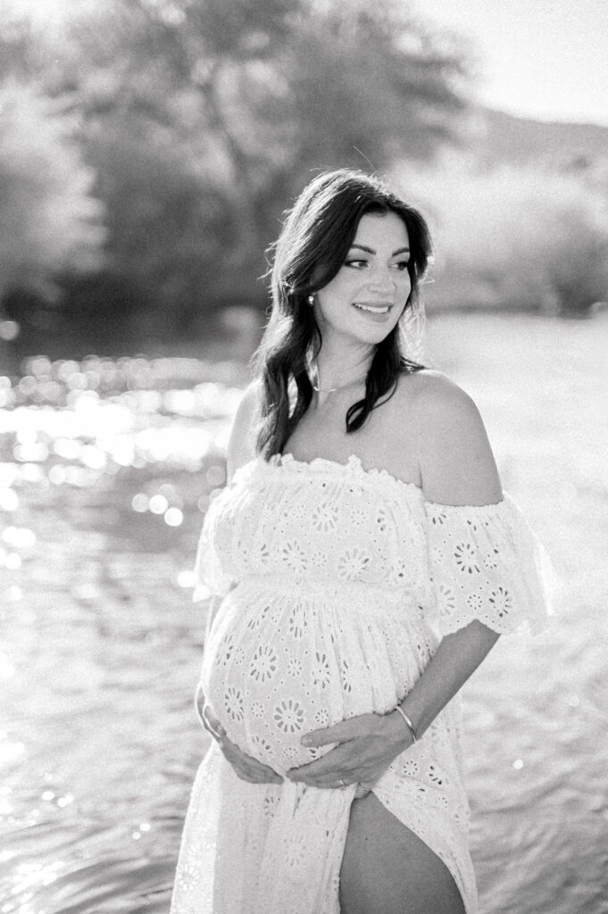 A pregnant woman in a white off-the-shoulder dress stands by the Salt River, smiling and holding her belly, with sunlight reflecting on the water—a serene Salt River Maternity moment.
